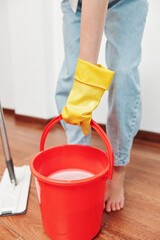 Woman with yellow rubber gloves holding a bucket of water on a hardwood floor with a mop in front of them