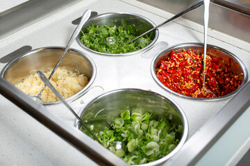 A view of a condiment bar, featuring containers for garlic, scallion, red chili, and cilantro, seen at a local restaurant.