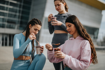 Three fit friends sharing a post-exercise moment with nutritious smoothies and snacks against an urban backdrop.