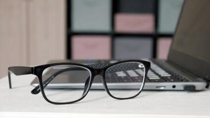 Close-up of black-framed glasses lying on a working table near a laptop