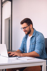 Fototapeta premium A smiling man with glasses is using a laptop in the office while sitting at his desk.