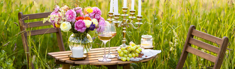 Beautiful romantic outdoor wedding decor in a field. Table decorated with burning candles, flowers. Wineglasses with white wine. Sunset, summer, golden hour. Perfect surprise date for couple. Banner