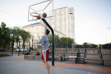 man in a grey hoodie and red shorts prepares to execute a basketball layup on an outdoor court. The basketball is about to enter the hoop