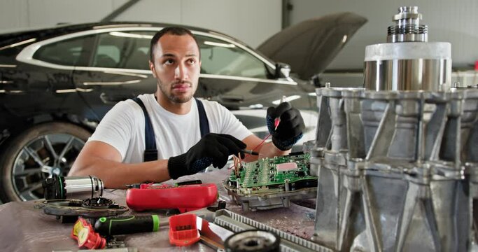 Male Auto Mechanic Inspecting Car Engine in Workshop. Mechanic working on an electric car's control unit in a workshop. The service includes voltmeter usage and intricate engine repairs