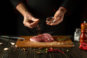 Chef sprinkles spices into meat steak for cooking for barbecue lunch. Working environment on the kitchen table with fragrant rosemary and garlic