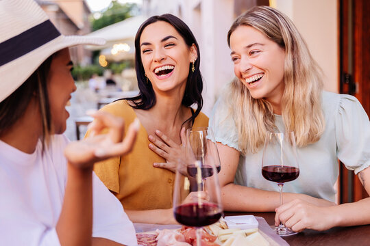 Three happy diverse women having fun enjoying aperitif with red wine sitting at an outside table in a bar. Food and beverage concept.