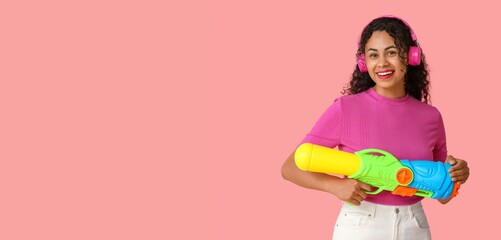 Happy African-American woman with water gun and headphones on pink background with space for text