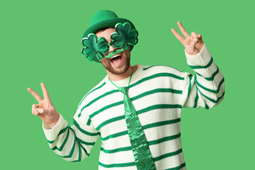 Happy young man in leprechaun's hat with party glasses showing victory gesture on green background. St. Patrick's Day celebration