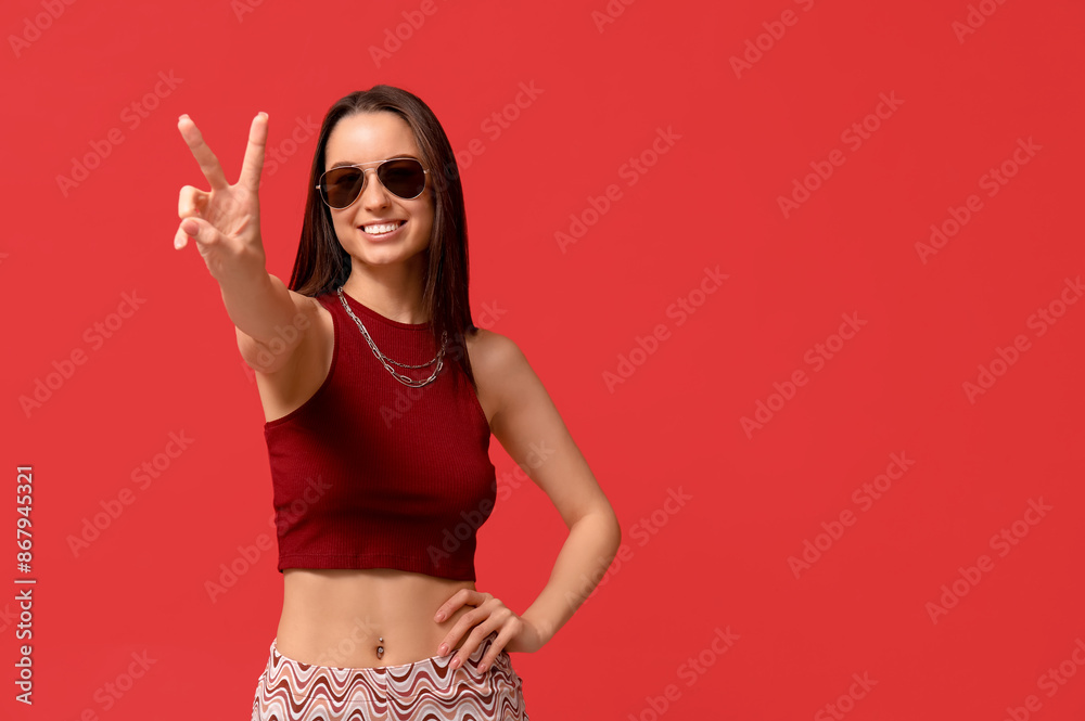 Wall mural Beautiful young happy woman with piercing navel showing victory gesture on red background