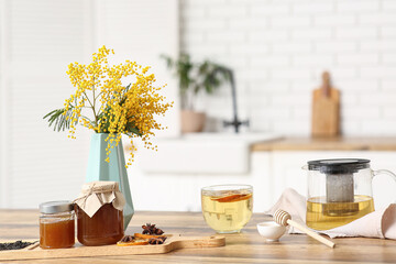 Teapot and cup of green tea with jar of honey and mimosa on table in kitchen