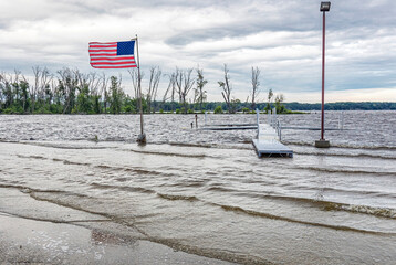 Fototapeta premium A river flooding over a road with a flooded pier and American flag on a windy, stormy day.