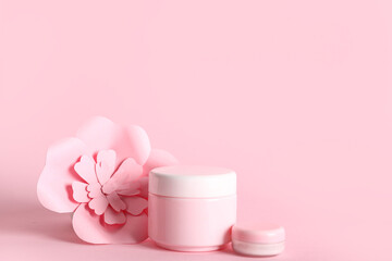 Jars of cosmetic cream with paper flowers on pink background