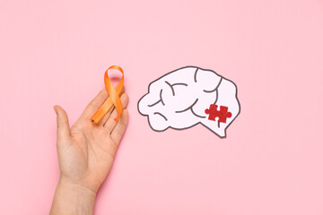 Female hand with orange ribbon, paper brain and puzzle piece on pink background. Multiple Sclerosis Awareness Month