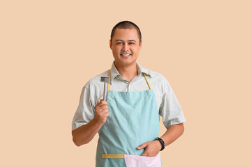 Young man in apron with meat tenderizer on beige background