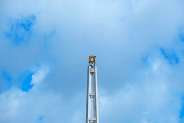 A tall, white synagogue spire featuring a prominent golden Star of David at its peak stands against a backdrop of a blue, cloud-filled sky.