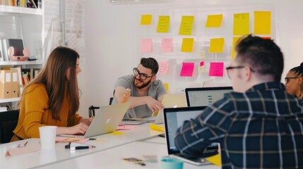 A group of young professionals are working together in a creative office space. They are using laptops and sticky notes to brainstorm ideas. The image is well-lit and has a positive atmosphere. It is