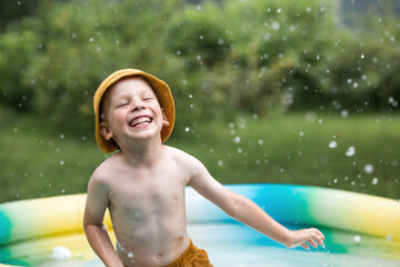 It's a hot day. A cute boy is swimming and splashing in the pool. A happy little boy smiles at the camera. Splashes of water are flying in all directions. The concept of summer holidays with children.