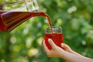 child hands holding glass and cherry juice pouring from jug with green garden on the background