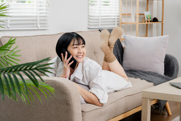 Young woman lying on a sofa, talking on the phone, in a cozy and modern living room with natural light and indoor plants.