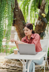 A young woman working on a laptop in an outdoor garden setting with a coffee cup, surrounded by greenery. She is wearing casual clothing and appears focused on her work.