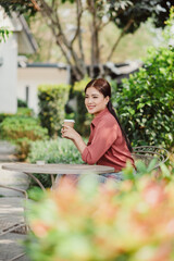 Naklejka premium A young woman in a red shirt enjoys a coffee outdoors, sitting at a table surrounded by lush greenery and trees.
