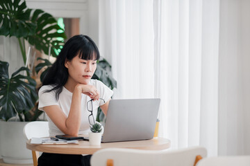 Young woman working on a laptop at home, sitting at a table with a plant and notebook, in a bright and modern room with natural light.