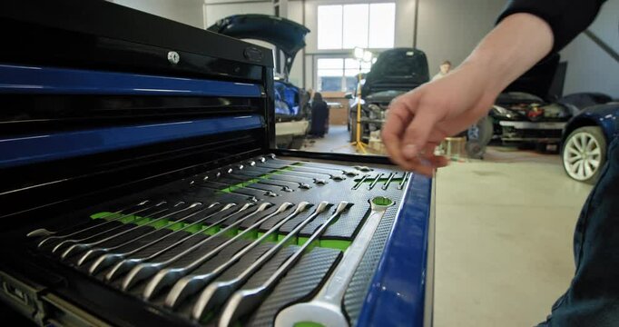 A mechanic handling tools and working on an electric car in a well-lit auto repair shop with blue tool cabinet. Mechanic Accessing Toolbox In Electric Car Repair Workshop.