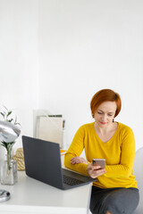 red haired woman in a yellow jacket sits with a laptop on a white table against a white wall. woman in business