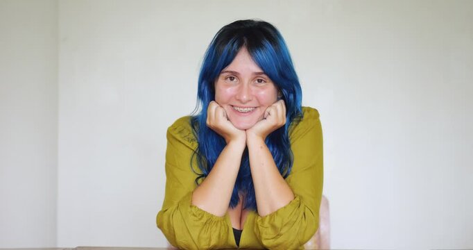 Portrait of positive confident woman with bright blue hair sits at table, smiling and radiating charm. Her elbows rest on table as she leans in, exuding sense of attentiveness and self-assuredness.