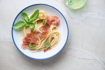 Linguine served with prosciutto and fresh basil in a blue and white plate, top view on a light-beige stone background, horizontal shot with space