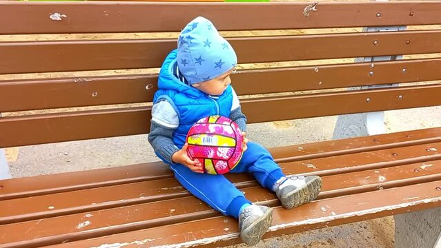 European boy one year old in cool weather sits on a bench in a blue vest with a pink ball