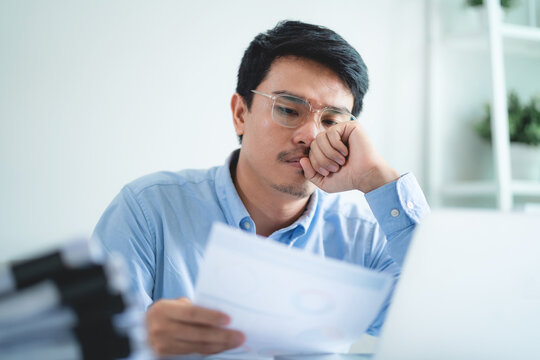 A man is sitting at a desk with a laptop and a stack of papers. He is looking at a piece of paper and he is in a state of deep thought