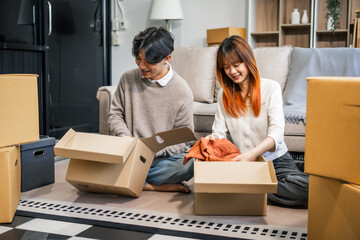 Indoors, a young couple happily packs cardboard boxes, preparing for a move. They share smiles and excitement, embracing new beginnings together in their new home, filled with love and anticipation