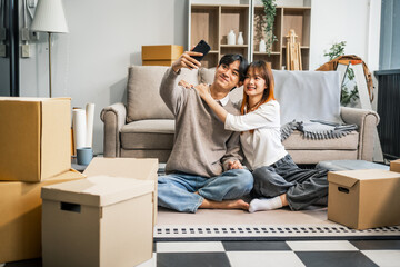  A young couple sits on the sofa, happily taking photos together and inspecting fingernail tabs while packing their belongings into a large box for their move to a new home. They share smiles