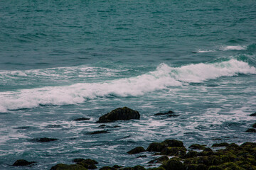 Large waves crash into with coastal rocks covered in green moss. Dramatic sea coast background. Raglan, New Zealand