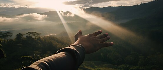 Hand reaching for sunbeams breaking through clouds over mountain landscape. Concept of hope, ambition, success and new beginnings.