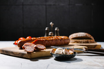 Smoked homemade sausage ring on the wooden cutting board with bowl of garlic, bread, tomato and knife. Rustic style. Hard light.