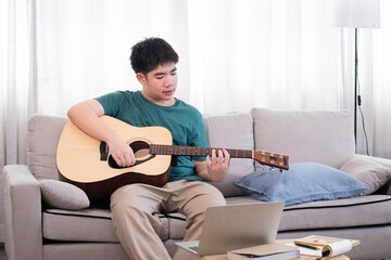 Young Asian male practicing guitar in a cozy living room setting, concentrating on learning chords with a laptop open for guidance. The casual, relaxed environment enhances his musical journey.