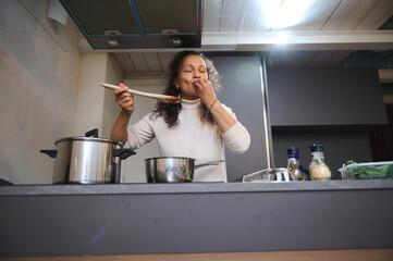 Young Italian woman tasting sauce on a wooden spoon, standing at kitchen countertop in modern minimalist home interior, showing sign of good taste, Bellissimo gesture, kissing fingers