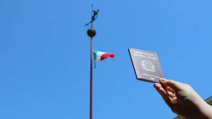 Hand holding an Italian passport with the Italian flag waving in the background under a blue sky