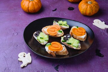 Sandwichs with cheese and cucumber in a Halloween decor in the form of mummies and pumpkins on a black oval plate on a purple concrete background. Halloween concept.