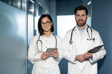 Fototapeta premium Healthcare professionals standing in hospital holding tablet and clipboard. Medical doctors wearing white lab coats and stethoscopes. Professional teamwork, healthcare service, hospital setting.
