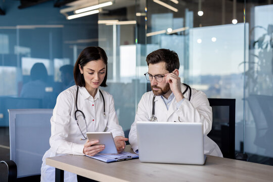 Medical professionals in lab coats collaborating in modern office environment using laptop and tablet for discussion and planning. Colleagues working together on healthcare project - Powered by Adobe