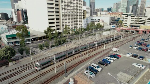 A modern train travels through a bustling city, surrounded by towering buildings and a parking lot.