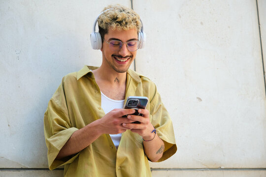 A man with a beard and glasses is smiling while holding a cell phone. He is wearing headphones and he is listening to music