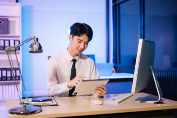 Obraz premium Overtime work concept, Handsome asian business man working overtime sitting at desk using computer and doing overtime project