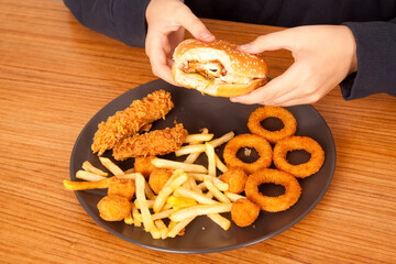Caucasian man eats half-eaten hamburger, potato chips, onion rings and chicken pieces in gray plate on wooden table