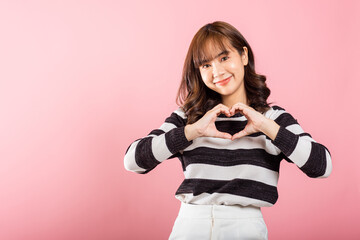 A confident woman smiles and creates a heart symbol with two hands on a pink background. Asian portrait of a beautiful young female expressing love and happiness for Valentine's Day.