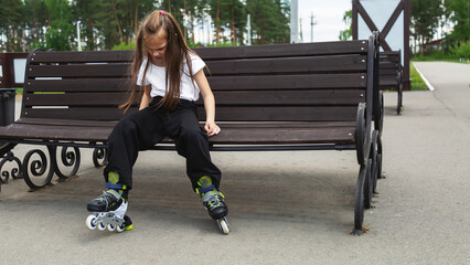 A little girl is skating in the park. A girl roller skates, sitting in a bench and putting on roller skates.