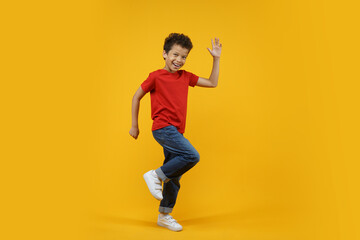 Full length studio portrait of beautiful happy smiling African American child dancing and waving with his hand, isolated over bright colored orange yellow background